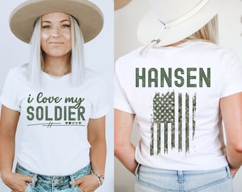 Könnte beinhalten: Weißes T-Shirt mit dem Text "i love my SOLDIER" auf der Vorderseite und dem Text "HANSEN" mit einer Tarnflagge der Vereinigten Staaten auf der Rückseite.