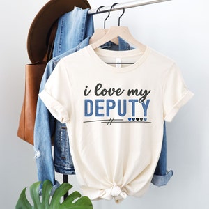 Pode incluir: Uma camiseta branca com o texto "i love my DEPUTY" em letras azuis e pretas. O texto &eacute; decorado com glitter azul. H&aacute; duas marcas de hashtag pretas e cinco cora&ccedil;&otilde;es abaixo do texto, dois azuis e tr&ecirc;s pretos.