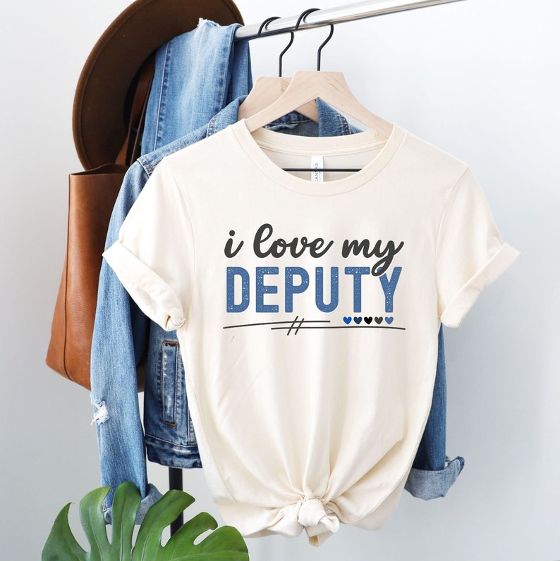 Pode incluir: Uma camiseta branca com o texto "i love my DEPUTY" em letras azuis e pretas. O texto &eacute; decorado com glitter azul. H&aacute; duas marcas de hashtag pretas e cinco cora&ccedil;&otilde;es abaixo do texto, dois azuis e tr&ecirc;s pretos.