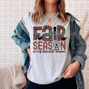May include: Gray t-shirt with a colorful graphic that says "Fair Season is my favorite season". The graphic includes a ferris wheel, a cow, a chicken, and a pig.