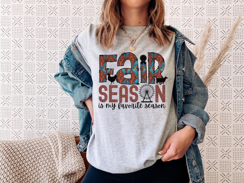 May include: Gray t-shirt with a colorful graphic that says "Fair Season is my favorite season". The graphic includes a ferris wheel, a cow, a chicken, and a pig.