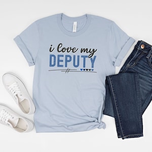 Pode incluir: Camiseta azul claro com o texto "i love my DEPUTY" em letras pretas e azuis. A camiseta est&aacute; colocada sobre uma superf&iacute;cie branca com um par de t&ecirc;nis brancos e uma cal&ccedil;a jeans azul.