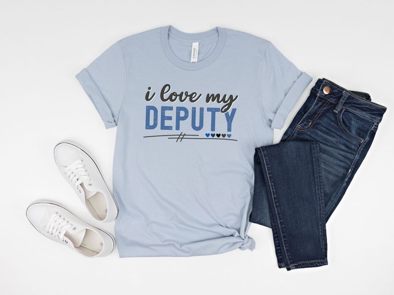 Pode incluir: Camiseta azul claro com o texto "i love my DEPUTY" em letras pretas e azuis. A camiseta est&aacute; colocada sobre uma superf&iacute;cie branca com um par de t&ecirc;nis brancos e uma cal&ccedil;a jeans azul.