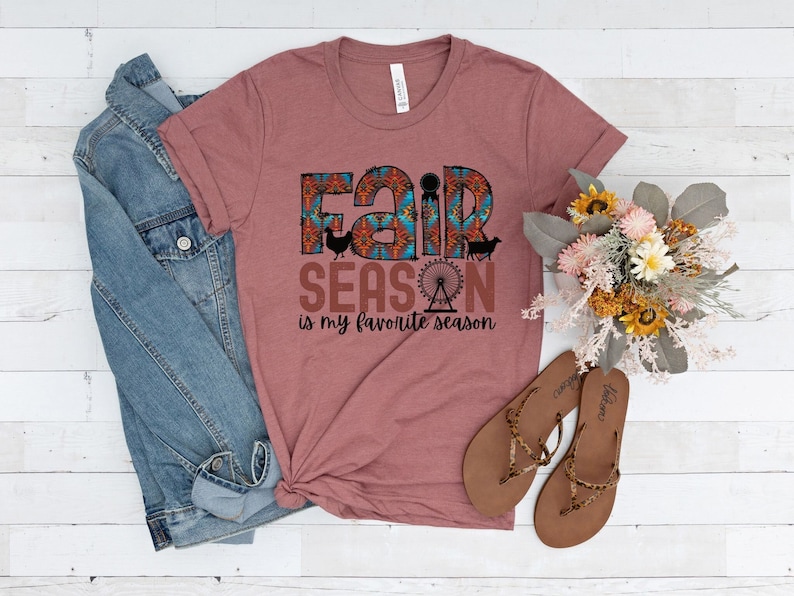 May include: A mauve t-shirt with a graphic print that says "Fair Season is my favorite season". The graphic features a colorful fairground design with a Ferris wheel, a chicken, a pig, and a goat.