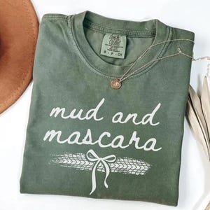May include: A green t-shirt with the text "mud and mascara" in white lettering. The shirt has a white tire track design with a white bow on top of the tire track.