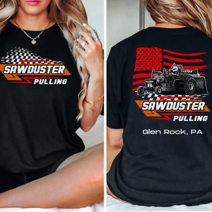 May include: Black t-shirt with a red, white, and blue American flag design on the back. The flag has a tractor pulling a sled in front of it. The text "Sawduster Pulling Glen Rock, PA" is printed below the tractor. The front of the shirt has the text "Sawduster Pulling" in white with a checkered flag design.