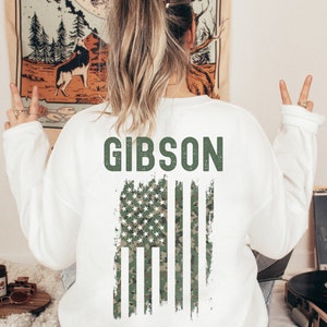 Pu&ograve; includere: Felpa bianca con un design di bandiera americana in mimetica verde e la parola "GIBSON" in testo verde.
