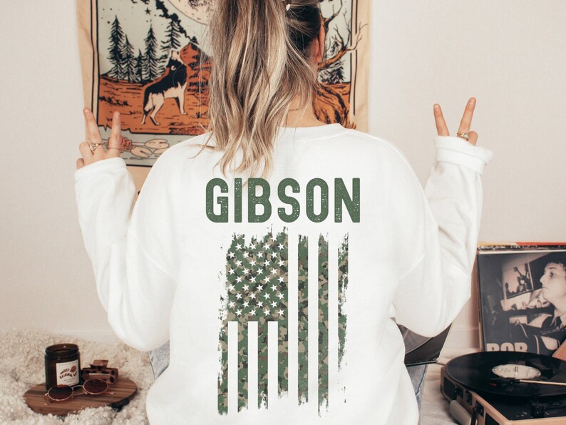 Pu&ograve; includere: Felpa bianca con un design di bandiera americana in mimetica verde e la parola "GIBSON" in testo verde.