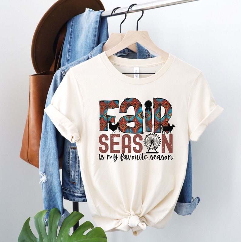 May include: A white t-shirt with a brown and blue graphic that says "Fair Season is my favorite season". The graphic includes a Ferris wheel, a chicken, and a cow.