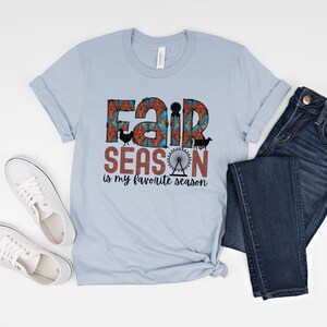 May include: A light blue t-shirt with a graphic design that says "Fair Season is my favorite season". The graphic features a colorful fairground design with a Ferris wheel, a cow, a chicken, and a pig.