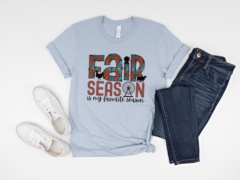 May include: A light blue t-shirt with a graphic design that says "Fair Season is my favorite season". The graphic features a colorful fairground design with a Ferris wheel, a cow, a chicken, and a pig.