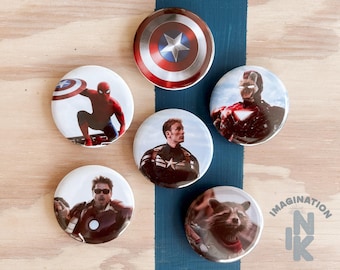 Peut inclure: Plusieurs boutons ronds et blancs représentant des personnages Marvel. Les boutons présentent le bouclier de Captain America, Spider-Man, Iron Man, Captain America et Rocket Raccoon. Ils sont disposés sur une surface en bois avec une bande bleue.