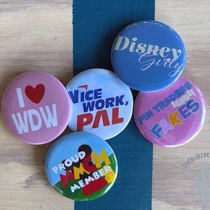 May include: Five circular pin-back buttons with various designs. One is pink with 'I heart WDW', one is blue with 'Disney Girly', one is white with 'Nice Work, Pal', one is pink with 'Pin Traders Against Fakes', and one is blue, yellow, and red with 'Proud MMCH Member'.
