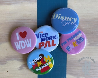 Peut inclure: Cinq badges ronds avec des motifs variés. Un rose avec 'I heart WDW', un bleu avec 'Disney Girly', un blanc avec 'Nice Work, Pal', un rose avec 'Pin Traders Against Fakes', et un bleu, jaune et rouge avec 'Proud MMCH Member'.