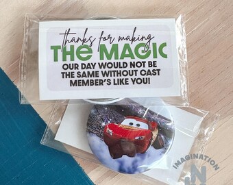 Peut inclure: Un badge rond avec un personnage de voiture de course rouge sur un fond bleu et blanc, emballé dans une pochette en plastique transparente avec une carte blanche portant l'inscription : Thanks for making THE MAGIC our day would not be the same without cast member's like you!