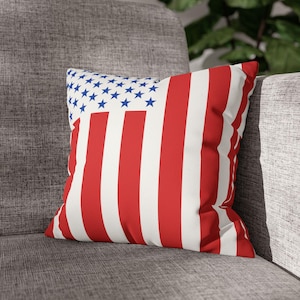 May include: Square throw pillow featuring the American flag design. The pillow has a white background with red stripes and a blue square with white stars in the upper left corner. The pillow is placed on a gray couch.