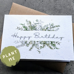 May include: A white card with a watercolour illustration of green leaves and white flowers. The card says "Happy Birthday" and has a green circle with the text "PLANT ME" on the bottom left corner.