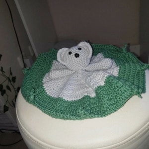 May include: A crocheted baby blanket with a white bear head in the center. The blanket is green and white and has a scalloped edge.