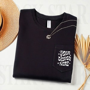 Op de afbeelding: Zwart T-shirt met een zakje aan de linkerkant. Het zakje heeft witte tekst die zegt "YOUR DESIGN HERE".