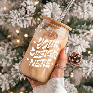 May include: A clear glass tumbler with a bamboo lid and a clear straw, filled with a creamy beverage. The tumbler has the text "YOUR DESIGN HERE" in white. The background is a blurred winter scene with lights and pinecones.