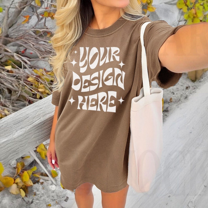 May include: A brown oversized t-shirt with the text "YOUR DESIGN HERE" in white. The shirt is being worn by a person with long blonde hair and a white tote bag.