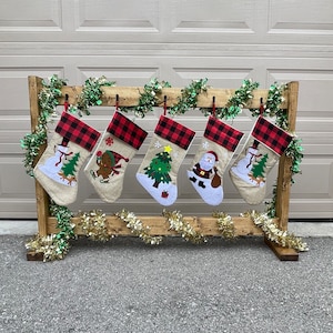 Puede incluir: Seis calcetines de Navidad de cuadros rojos y blancos con diferentes diseños bordados, incluyendo un muñeco de nieve, un hombre de jengibre, un árbol de Navidad, Papá Noel y un muñeco de nieve con un reno. Los calcetines están colgados en un estante de madera con una guirnalda verde.