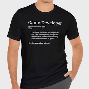 Może przedstawiać: Czarny T-shirt z białym tekstem, który brzmi "Game Developer [geym dih-vel-uh-per] noun 1. A digital illusionist, turning coffee into code and bugs into unexpected features. Can summon a functioning game from the chaos of syntax... See also: magician, wizard"