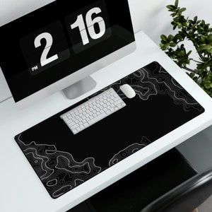 Puede incluir: Una alfombrilla de escritorio negra y blanca con un diseño topográfico. Un teclado y un ratón blancos están sobre la alfombrilla. La alfombrilla está sobre un escritorio blanco con un monitor de ordenador en el fondo.