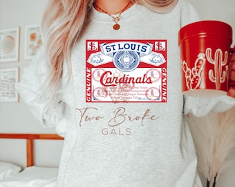 May include: A gray sweatshirt with a vintage-style St. Louis Cardinals logo printed on the front. The logo is red, white, and blue and features the team's name and the words "Genuine" and "Genuine". Below the logo is the text "Two Broke Gals".