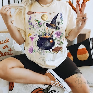 May include: Cream-colored t-shirt featuring a whimsical witch-themed design. The graphic includes a witch's hat, cauldron, frog, broom, moon, and flowers. The text "Two Broke Gals" is printed at the bottom of the shirt.