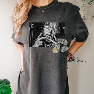 May include: A black and white photo of a man wearing a camouflage hat and a gold chain. The photo is printed on a gray t-shirt with the text "Two Broke Gals" printed below it.