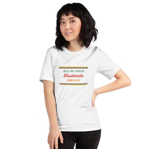 May include: A white short-sleeved t-shirt featuring a graphic design with horizontal stripes in yellow, orange, and green. The text reads ALL OF YOUR in green, Husbands in red cursive script, and ARE GAY in orange, centered between the stripes.