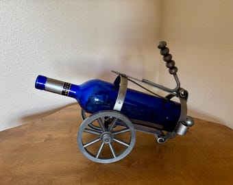 May include: A decorative wine bottle holder shaped like a miniature wagon. The holder is made of metal and features a blue glass bottle. The wagon has spoked wheels and a handle with wooden beads.