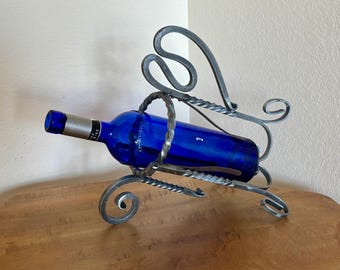 May include: A handcrafted metal wine bottle holder with a decorative design. The holder cradles a cobalt blue wine bottle. The metalwork features swirling accents and a twisted handle, adding a touch of elegance to the piece.