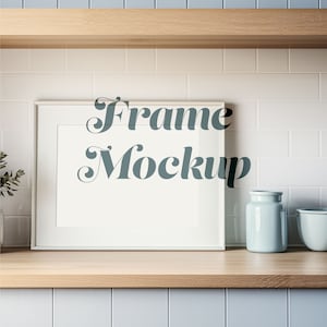 May include: White picture frame mockup on a wooden shelf with a white tile backsplash. The frame is empty and ready for your design.