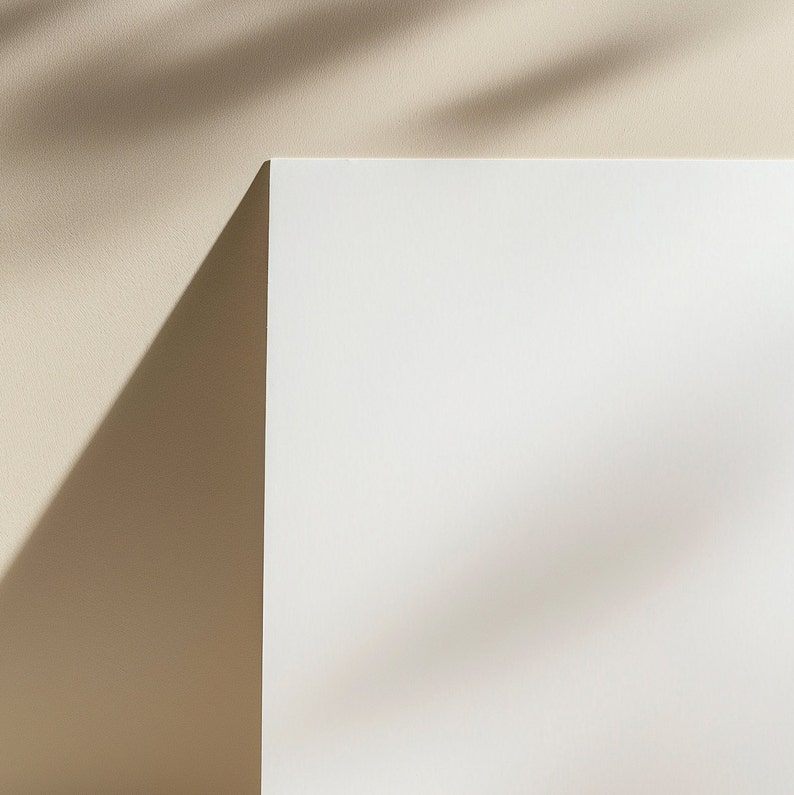 May include: A white piece of paper sits on a light brown surface. The paper is blank and has shadows cast on it from the light above.