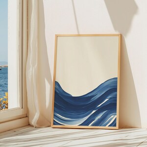 May include: A framed abstract painting of blue waves on a cream background. The painting is in a light wood frame and is leaning against a white wall.