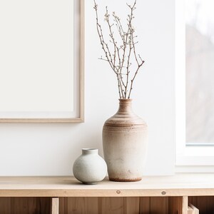 May include: Two ceramic vases, one light brown and one light green, on a wooden shelf. The brown vase is taller and holds dried branches with white blossoms.