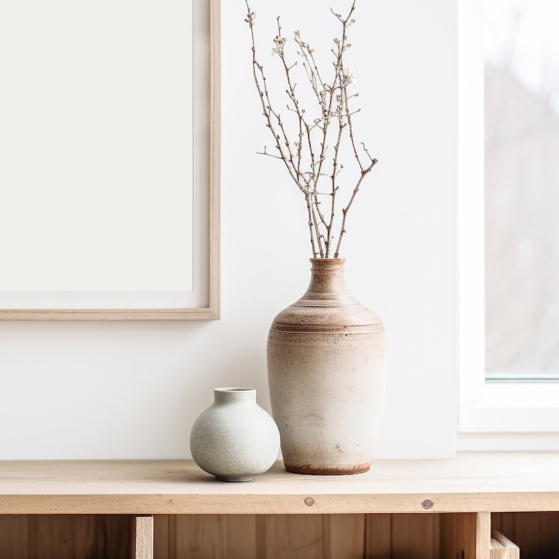 May include: Two ceramic vases, one light brown and one light green, on a wooden shelf. The brown vase is taller and holds dried branches with white blossoms.