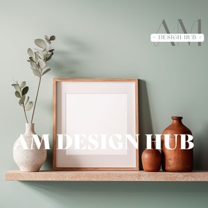 May include: A wooden frame with a white mat and a white background sits on a shelf with a white vase with green leaves, a small brown ceramic vase, and a larger brown ceramic vase. The text "AM DESIGN HUB" is printed in white on the shelf.