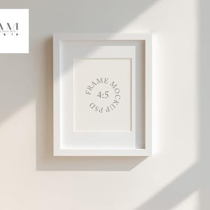 May include: A white rectangular picture frame with a white mat, hanging on a white wall. The frame contains the text "FRAME MOCKUP 4:5 PSD" in a circular design. The image is lit by natural light, casting shadows.
