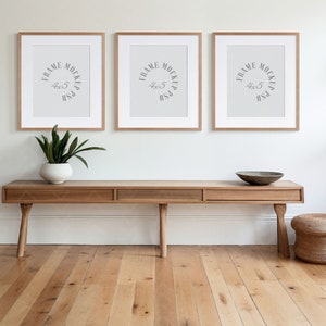 May include: Three framed art mockups with the text "FRAME MOCKUP 4x5 PSD" are displayed above a wooden console table. A white vase with a green plant and a decorative bowl are on the table. A woven stool sits to the right.