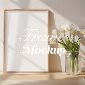 May include: A wooden frame mockup with a white background. The frame is sitting on a light wood surface. A bouquet of white tulips in a clear glass vase is to the right of the frame.