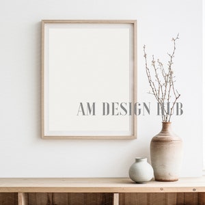 May include: A wooden frame with a white background sits on a wooden shelf. A vase with a light brown and white glaze holds a bouquet of dried branches with small white flowers. A smaller, light blue vase sits next to the larger vase.