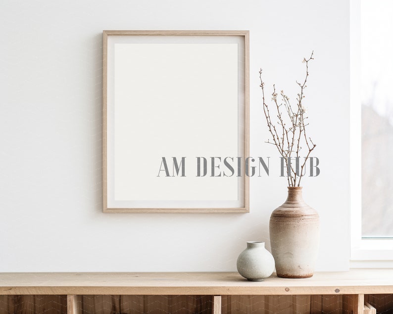 May include: A wooden frame with a white background sits on a wooden shelf. A vase with a light brown and white glaze holds a bouquet of dried branches with small white flowers. A smaller, light blue vase sits next to the larger vase.