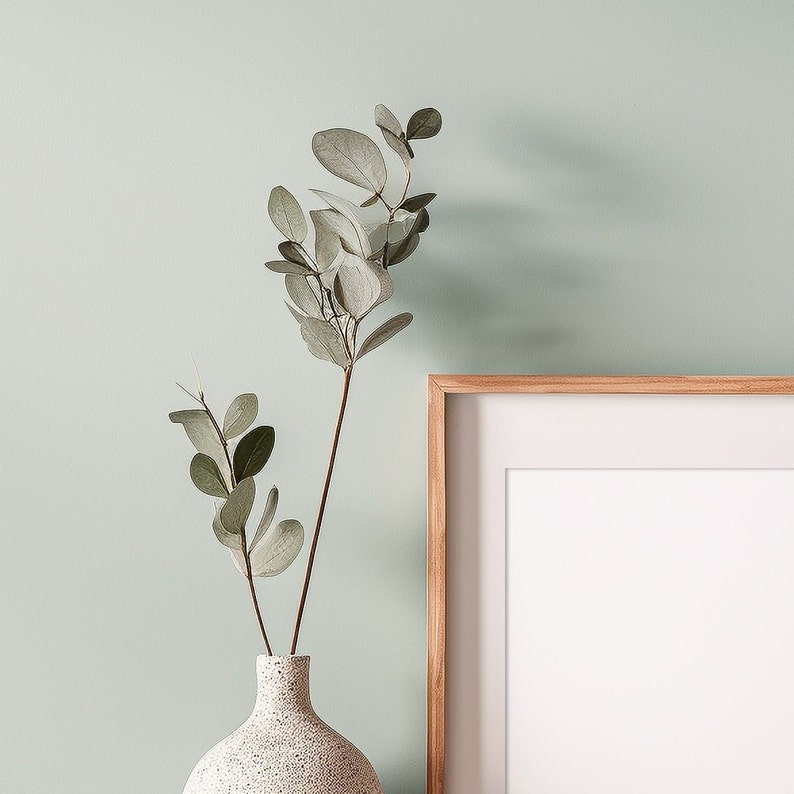 May include: A light green wall with a wooden frame picture and a vase with a sprig of eucalyptus.