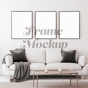 May include: Three black picture frames with white mats are hanging on a white wall above a white couch with gray throw pillows and a gray throw blanket. The couch is in front of a coffee table with two white mugs on it.