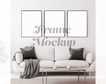 May include: Three black picture frames with white mats are hanging on a white wall above a white couch with gray throw pillows and a gray throw blanket. The couch is in front of a coffee table with two white mugs on it.