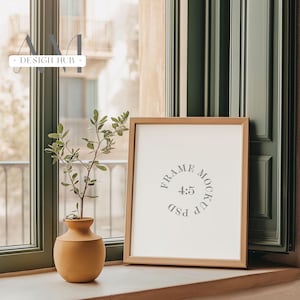 May include: A light brown wooden frame with a white mat and a white background. The frame is sitting on a windowsill with a small potted plant in front of it. The frame has the text "FRAME MOCKUP 4:5 PSD" printed on the white background.