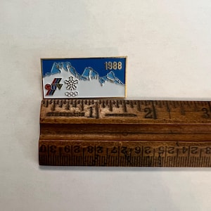 May include: A rectangular pin with a gold border. The pin features a blue background with white mountain silhouettes and the year "1988". The Olympic rings and a snowflake design are also present. The pin is approximately 1 inch long.